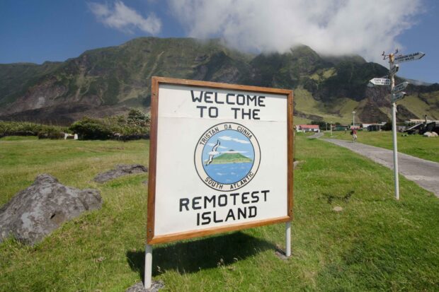 A road sign reading "Welcome to the remotest island," mountains are visible in the background.
