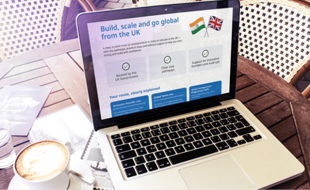 A laptop displaying a webpage titled "Build, scale and go global from the UK" with UK and Indian flag. Beneath the title is the text, “A clear, trusted route for entrepreneurs in India to relocate to the UK, highlighting support for entrepreneurs through UK government backing, clear visa pathways, and innovation funding. There is a cup of coffee and some papers on the table with the laptop.