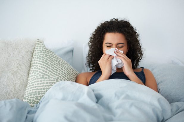 A woman sitting in bed under a duvet blowing her nose