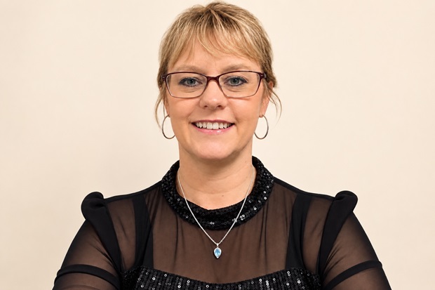 A picture of a smiling lady wearing glasses, hair tied back and brown blouse.