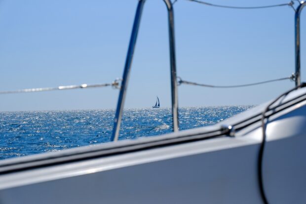 A sailing vessel in the distance on a blue sea framed through a railing of another boat