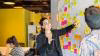 A young woman inside pointing to a whiteboard with colourful stick-on notes, explaining work processes to another male colleague in the office