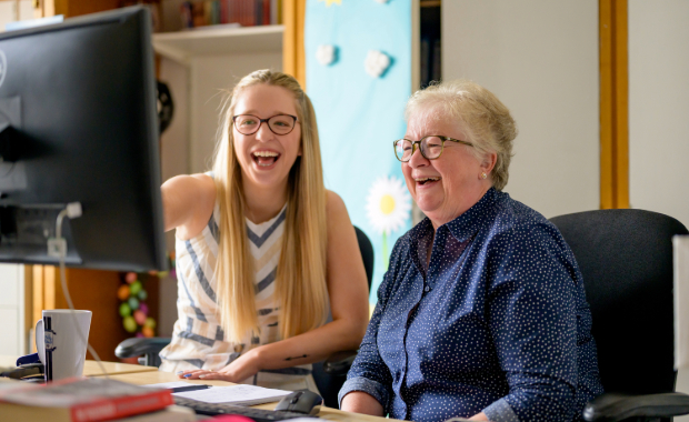A couple of women, one young, the other middle-aged are sat in front of a computer. They are laughing at something on the screen.
