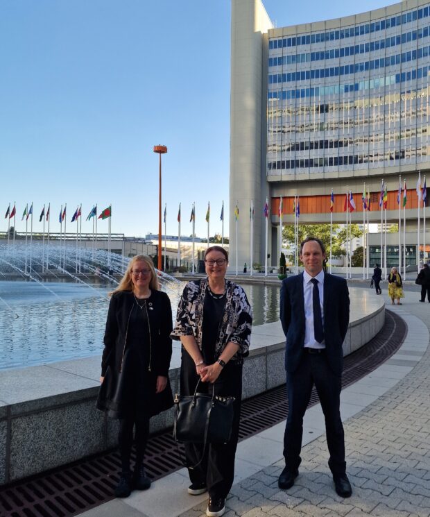 Jo Nettleton standing with two colleagues beside fountains and international flags outside the International Atomic Energy Agency headquarters in Vienna.