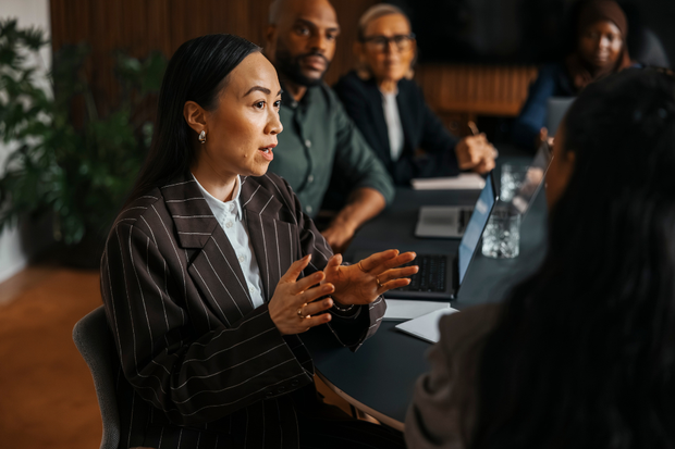 A professional woman is talking to colleagues in a boardroom, gesturing with her hands.