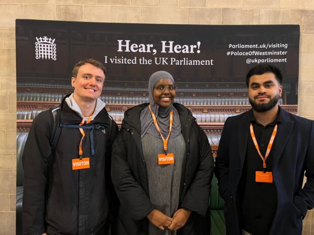 Three young people, two male, one female stood infront of a banner detailing their location at the Houses of Parliament.