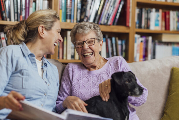 Image of an elderly mother and her daughter, smiling whilst reading a book on the sofa.