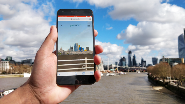 A hand holding a phone depicting the VU.CITY app with a backdrop of the river thames and London skyline.