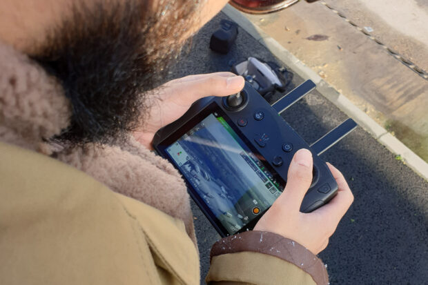 Close-up of a person holding a drone controller with a built-in screen displaying a live aerial view of HGV lorries in a large depot. This drone footage was featured in a video project for the Department for Transport.