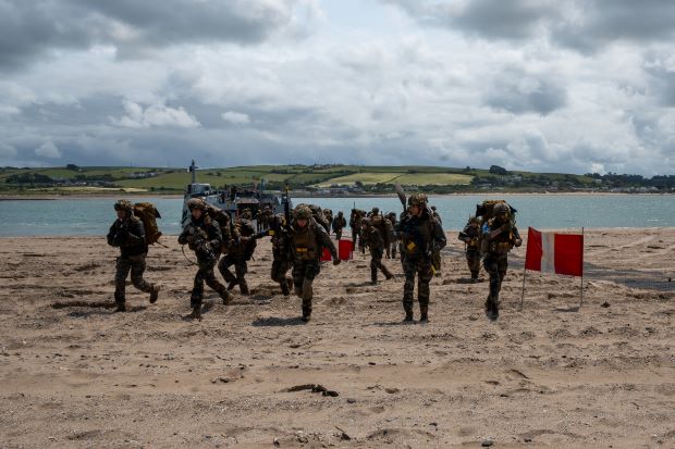 A group of military personnel with large backpacks and weapons run towards the camera on a beach.