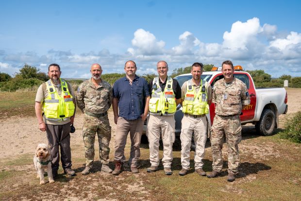 A group of men in military clothing and high vis jackets smile to the camera on a military training area