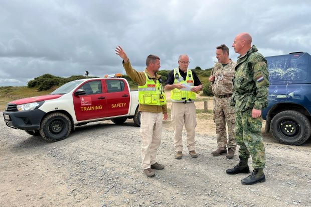 Four people, some in military uniform, some in high vis jackets, stand in front of a military range car on a training area.