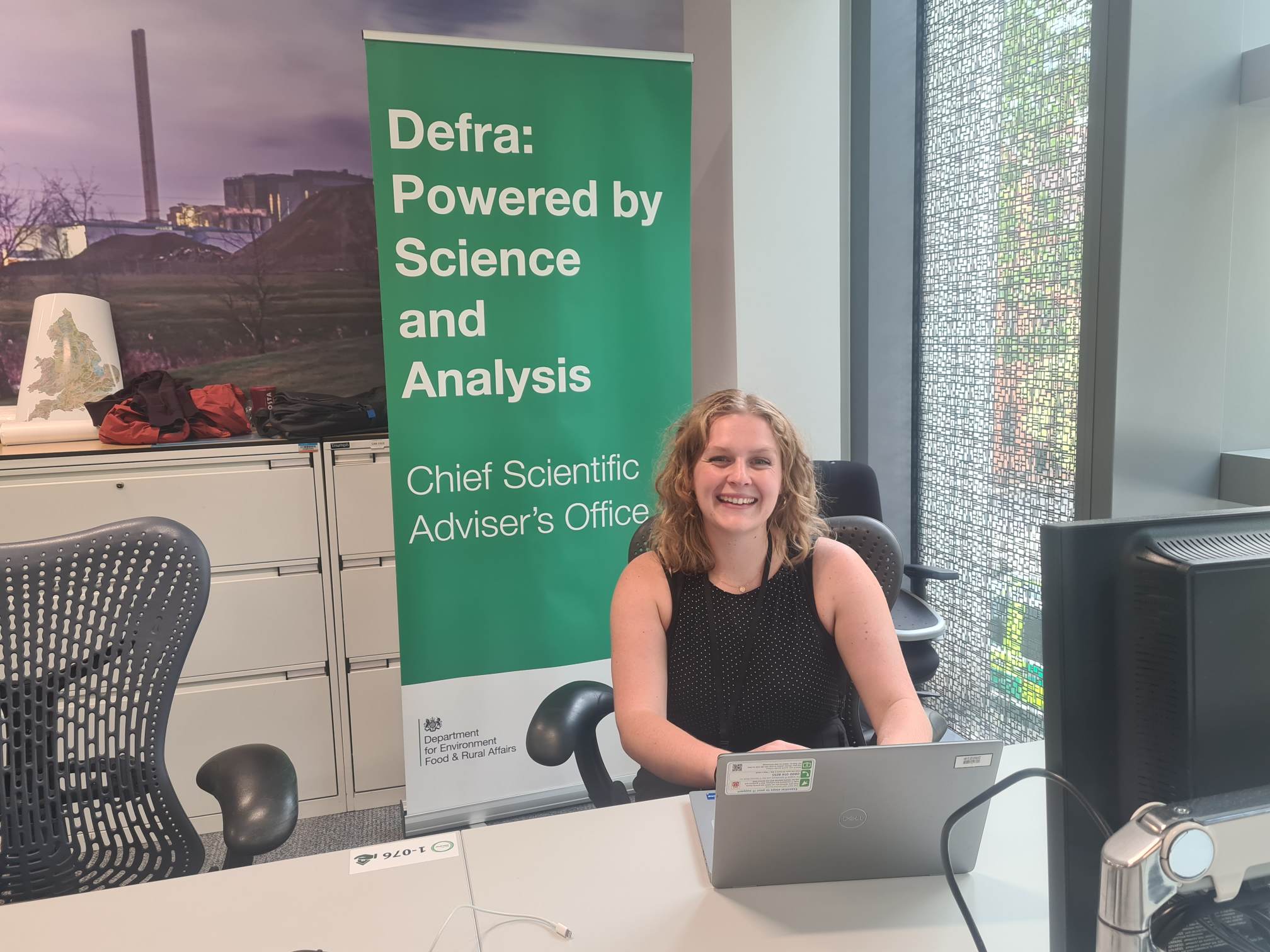 A photo of Sadie Sweetland sitting at a desk with a laptop, smiling. Behind her is a green banner that reads "Defra: Powered by Science and Analysis. Chief Scientific Adviserโs Office."