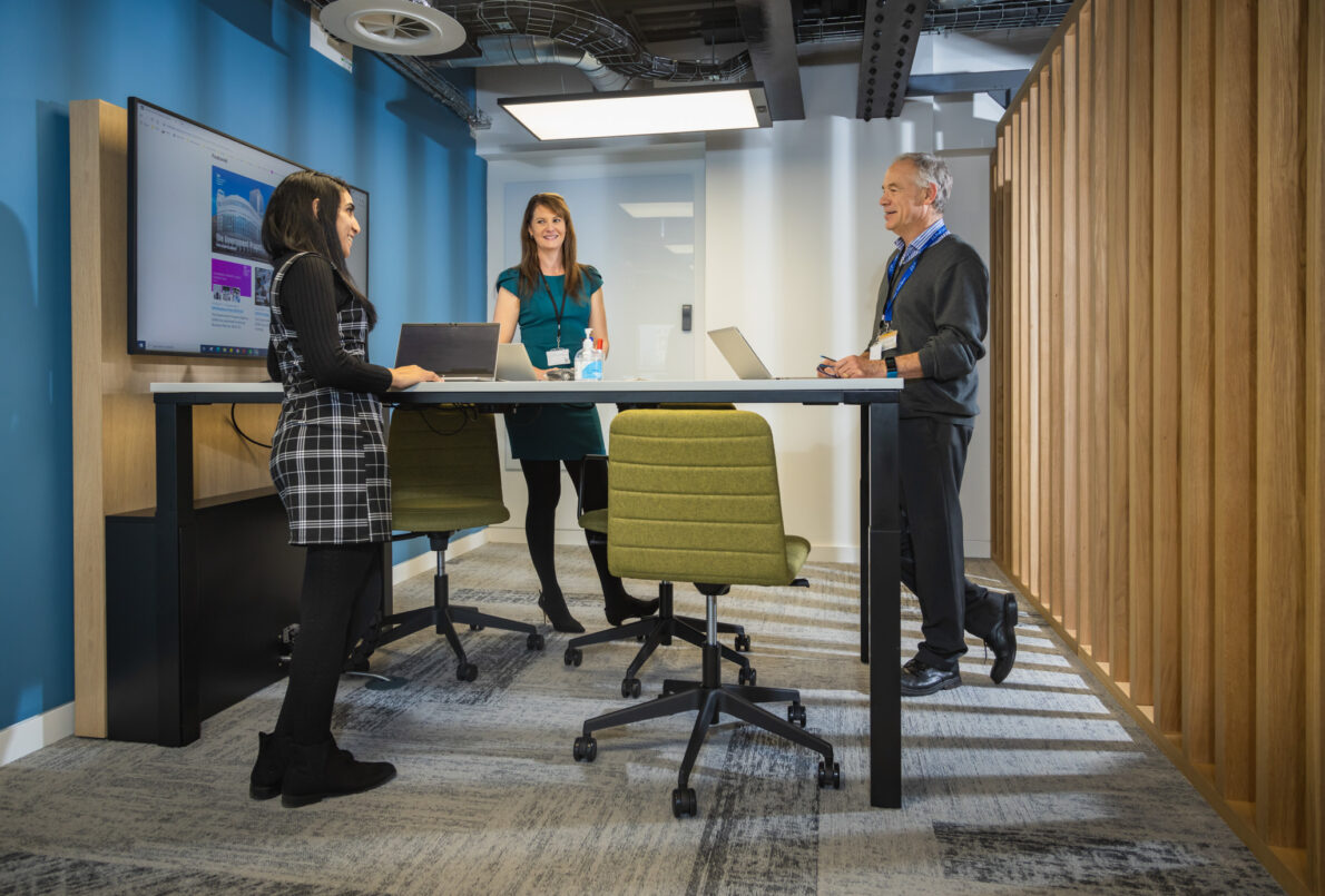 Flexible working space, 3 colleagues standing around a desk in a workspace with laptops on the table.