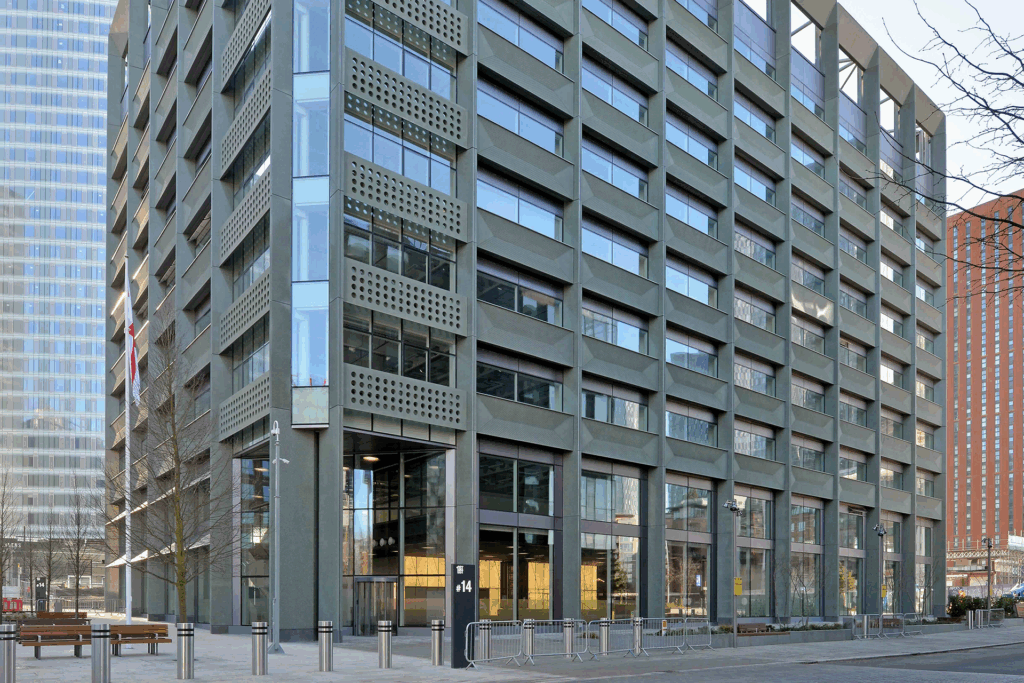 An exterior, close-up view of an eight-story modern office building in Manchester, England, featuring a dark green/grey perforated metal facade and large horizontal rectangular windows. The ground floor has a recessed glass entrance and the building is set back from a paved public space with bollards and benches.