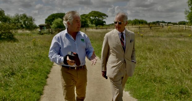 Tony walks with King Charles along a path in the Lincolnshire NNR. It's a beautiful sunny day, the King wears a light beige coloured suit. Tony has a blue shirt - he carries a pair of binoculars in his hand.
