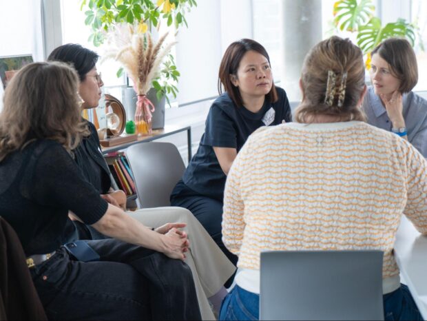 Five women of different ethnicities sitting while in a discussion in a modern office setting with a bookshelf and plants behind them.
